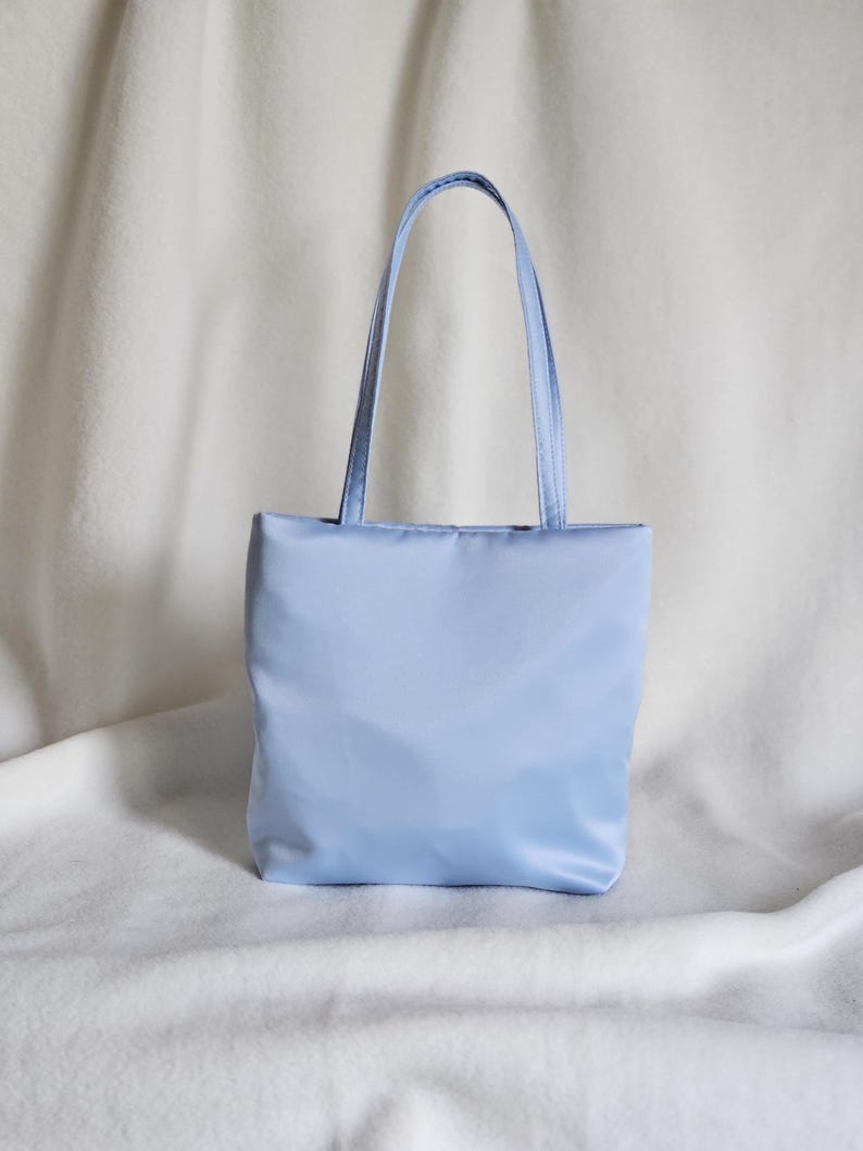 May include: A light blue tote bag with a smooth, solid-colored fabric exterior. The bag features a rectangular body and two matching light blue shoulder straps attached to the top edge, providing a simple and functional design for carrying items.