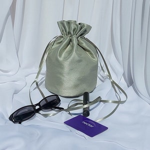 A round bottom dolly bag made from soft sheen faux silk fabric with ribbon drawstrings to fasten it. It is sage green coloured and is shown with a display of possible contents to show what will fit into the bag.