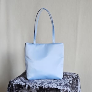 May include: A light blue tote bag with a rectangular body and two matching thin shoulder straps. The bag features a smooth, solid-colored fabric construction with a clean, vertical orientation and a simple, unadorned design throughout.