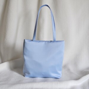 May include: A light blue tote bag with a rectangular body and two matching shoulder straps. The bag features a smooth, solid-colored exterior with a clean, structured design and a flat base, providing a simple and functional carryall accessory.