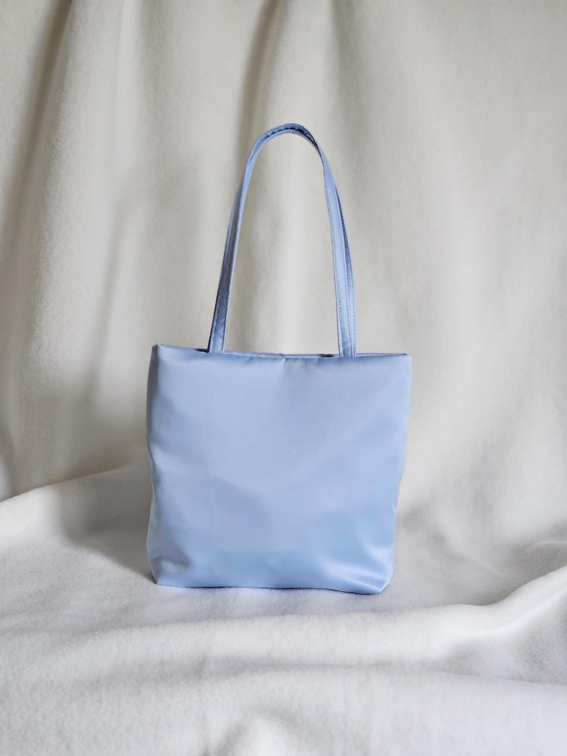 May include: A light blue tote bag with a rectangular body and two matching shoulder straps. The bag features a smooth, solid-colored exterior with a clean, structured design and a flat base, providing a simple and functional carryall accessory.
