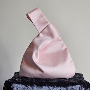 May include: A light pink satin handbag with a knotted handle. The bag has a triangular shape and a smooth, shiny texture. The handle is a loop of the same satin material. The bag is sitting on a dark blue velvet surface.
