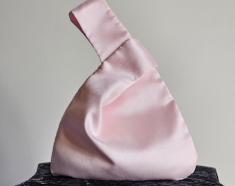 May include: A light pink satin handbag with a knotted handle. The bag has a triangular shape and a smooth, shiny texture. The handle is a loop of the same satin material. The bag is sitting on a dark blue velvet surface.