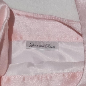 May include: Close-up of a light pink satin garment with a white lining. The label reads "Grace and Roses" in a delicate script. The fabric has a subtle sheen, suggesting a formal or delicate item.