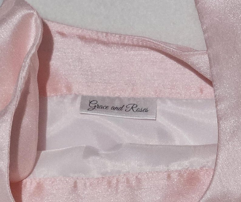May include: Close-up of a light pink satin garment with a white lining. The label reads "Grace and Roses" in a delicate script. The fabric has a subtle sheen, suggesting a formal or delicate item.
