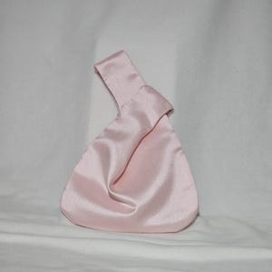 May include: A light pink satin knot bag with a handle. The bag has a triangular shape with a rounded bottom. The fabric has a subtle sheen, and the handle is formed by a knot at the top. The bag is on a white background.