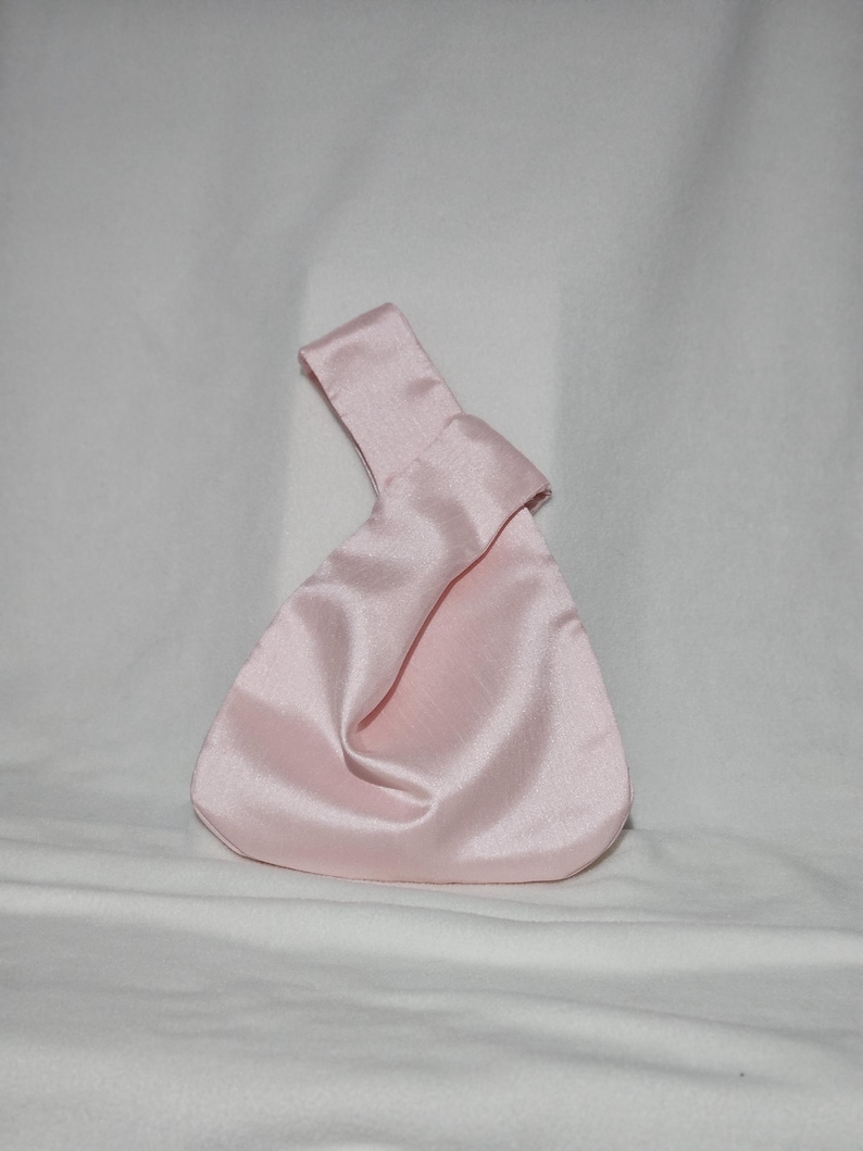May include: A light pink satin knot bag with a handle. The bag has a triangular shape with a rounded bottom. The fabric has a subtle sheen, and the handle is formed by a knot at the top. The bag is on a white background.