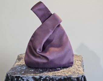 May include: A plum-colored satin knot bag. The bag has a triangular shape with a knotted handle. The fabric has a subtle sheen, and the bag sits on a dark, textured surface. The background is a plain, light color.