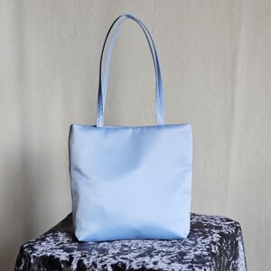 May include: A light blue tote bag with a smooth, solid-colored exterior and two matching vertical shoulder straps. The bag features a rectangular shape with a flat base and clean, minimalist stitching along the top and side edges of the main body.