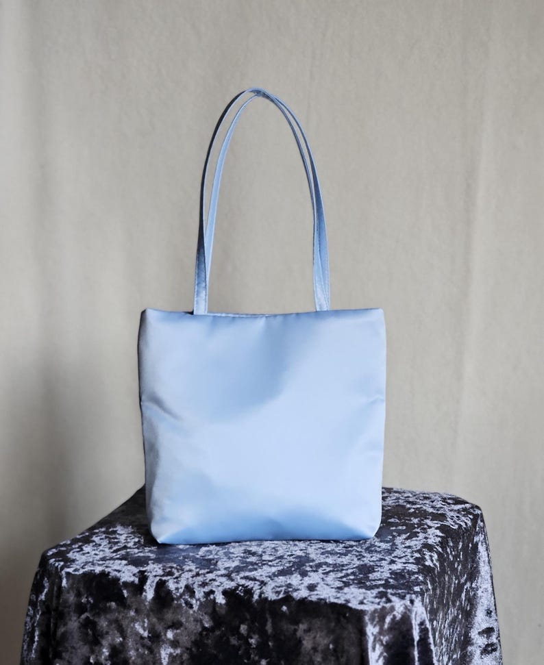 May include: A light blue tote bag with a smooth, solid-colored exterior and two matching vertical shoulder straps. The bag features a rectangular shape with a flat base and clean, minimalist stitching along the top and side edges of the main body.