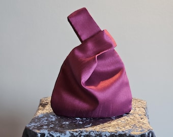 May include: A plum-colored satin knot bag with a handle. The bag is sitting on a dark gray velvet-covered surface. The bag's fabric has a slight sheen, and the handle is folded over the top.