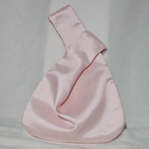 May include: A light pink satin knot bag with a handle. The bag has a triangular shape with a rounded bottom. The handle is a folded strip of the same satin material. The bag is made of a shiny, smooth fabric.