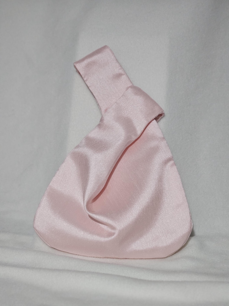 May include: A light pink satin knot bag with a handle. The bag has a triangular shape with a rounded bottom. The handle is a folded strip of the same satin material. The bag is made of a shiny, smooth fabric.