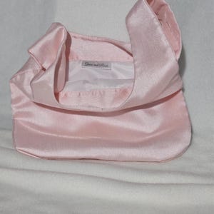 May include: A light pink satin handbag with a rounded bottom and a handle that loops over the top. The interior is white, and the label reads "Grace and Rose". The bag is sitting on a white surface.