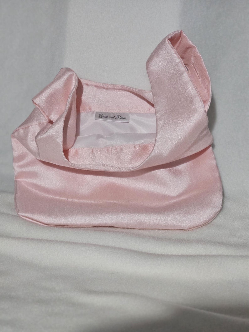 May include: A light pink satin handbag with a rounded bottom and a handle that loops over the top. The interior is white, and the label reads "Grace and Rose". The bag is sitting on a white surface.