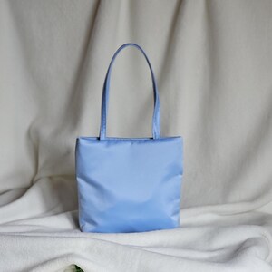 May include: A light blue rectangular tote bag with a smooth, solid-colored fabric exterior and two matching vertical shoulder straps. The bag features a clean, minimalist design with a flat base and a simple, unadorned front panel construction.