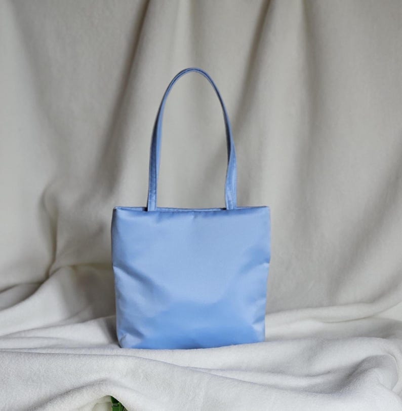 May include: A light blue rectangular tote bag with a smooth, solid-colored fabric exterior and two matching vertical shoulder straps. The bag features a clean, minimalist design with a flat base and a simple, unadorned front panel construction.