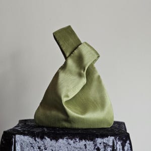 May include: A sage green satin handbag with a knotted handle. The bag has a triangular shape and sits on a black velvet surface. The fabric has a subtle sheen, and the handle is folded over the top.