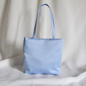 May include: A light blue tote bag with a smooth, satin-like finish. The bag features a rectangular body and two matching light blue shoulder straps attached to the top edge. The material has a subtle sheen, and the design is simple and structured.
