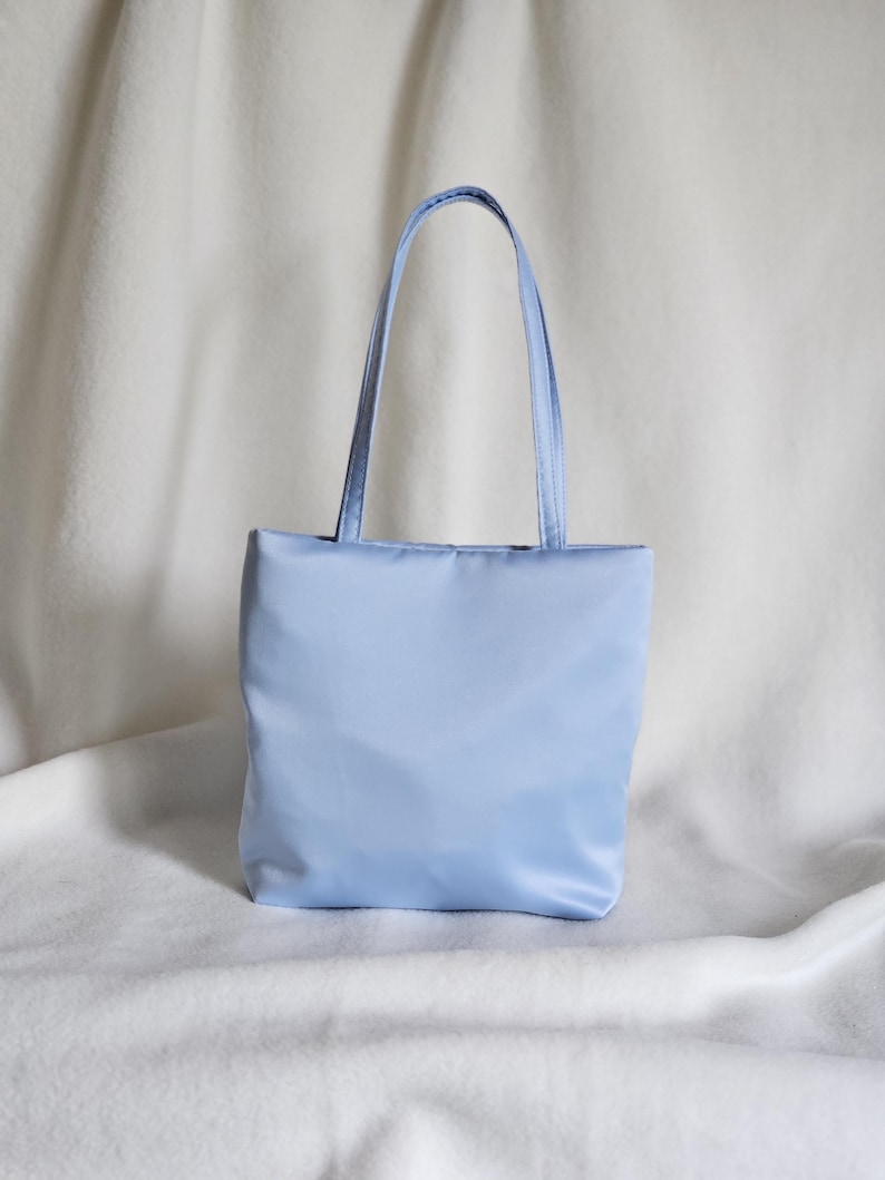May include: A light blue tote bag with a smooth, satin-like finish. The bag features a rectangular body and two matching light blue shoulder straps attached to the top edge. The material has a subtle sheen, and the design is simple and structured.