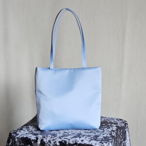 May include: A light blue tote bag with a rectangular body and two matching shoulder straps. The bag features a smooth, solid-colored exterior surface with a clean, vertical orientation and a simple, unadorned design throughout the entire piece.