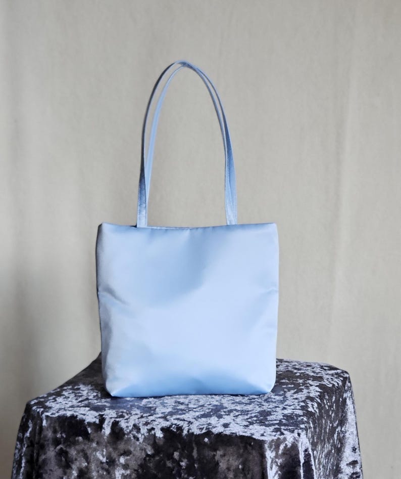 May include: A light blue tote bag with a rectangular body and two matching shoulder straps. The bag features a smooth, solid-colored exterior surface with a clean, vertical orientation and a simple, unadorned design throughout the entire piece.