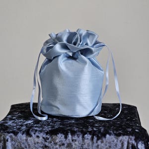 Könnte beinhalten: Ein hellblaues Satin-Kordelzugbeutel. Der Beutel ist oben mit einem Bandverschluss gerafft. Der Beutel steht auf einer dunkelblauen Samtoberfläche. Der Beutel hat ein weiches, elegantes Design.