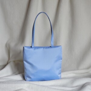 May include: A light blue tote bag with a smooth, solid-colored fabric exterior. The bag features a rectangular body and two matching light blue shoulder straps attached to the top edge, providing a simple and functional design for carrying items.