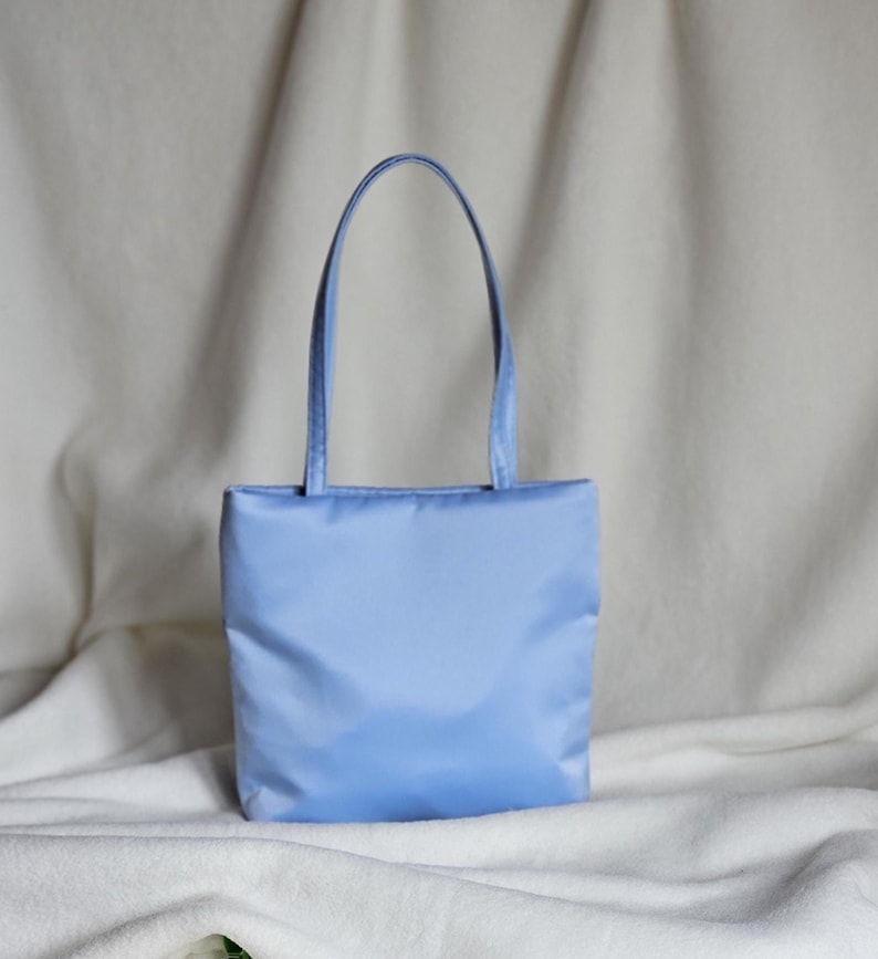 May include: A light blue tote bag with a smooth, solid-colored fabric exterior. The bag features a rectangular body and two matching light blue shoulder straps attached to the top edge, providing a simple and functional design for carrying items.
