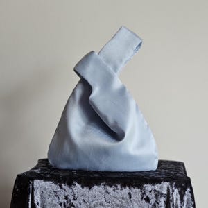 Könnte beinhalten: Hellblaue Satin-Handtasche mit geknotetem Henkel. Die Tasche hat eine dreieckige Form und steht auf einer schwarzen Samtoberfläche. Das Satinmaterial hat einen dezenten Glanz, der ihm ein luxuriöses Aussehen verleiht.