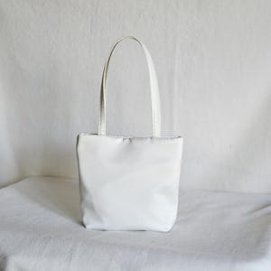 Può includere: Una borsa tote bianca con un corpo rettangolare e due manici abbinati. La borsa è realizzata in un materiale liscio, forse simile al raso. Lo sfondo è una superficie bianca.