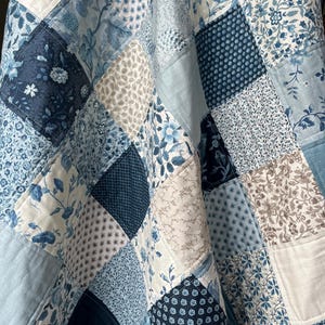 May include: A patchwork quilt in shades of blue and white, with floral and geometric patterns. The quilt is constructed from squares and rectangles of various fabrics, creating a textured design. The colour scheme includes light blue, navy, and cream, offering a classic aesthetic.