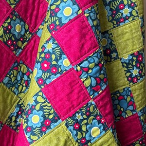 May include: A patchwork quilt with squares of pink, green and blue fabric featuring floral patterns. The quilt is folded in half and the fabric has a textured feel.