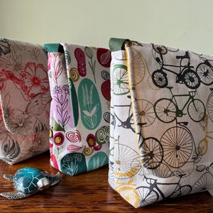 May include: Three fabric bags with different patterns. The first bag has a floral pattern in shades of pink, red, and green. The second bag has a pattern of swirls and flowers in shades of pink, green, and white. The third bag has a pattern of bicycles in black and white on a cream background.