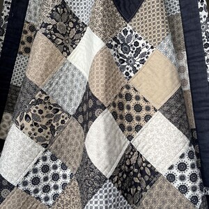 May include: A patchwork quilt featuring a repeating diamond pattern composed of square fabric blocks. The quilt displays a colour palette of black, white, cream, and tan, with various floral, geometric, and botanical prints on each individual square.
