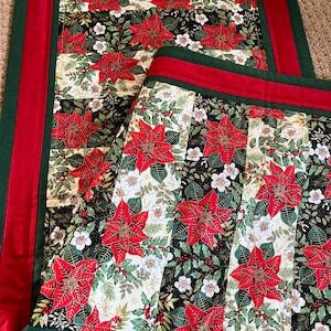 May include: A festive table runner and placemat set. The design showcases red poinsettias, white flowers, and green leaves against black and cream stripes. The edges are trimmed with red and green fabric, creating a seasonal look.