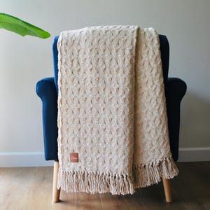 Puede incluir: Una manta de punto beige con un patrón texturizado y flecos está sobre un sillón azul. La manta tiene una etiqueta rectangular de cuero. La silla tiene patas de madera.