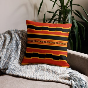 May include: Square throw pillow with a geometric pattern in red, black, and gold. The design features horizontal stripes and stepped shapes. The pillow is placed on a gray sofa with a blue and white patterned blanket.