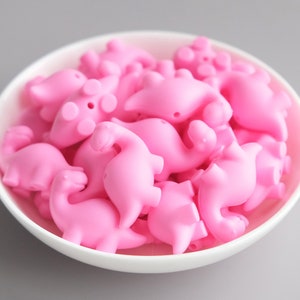 May include: A bowl filled with pink silicone dinosaur-shaped beads. The beads are all the same size and shape, and they are arranged in a random pattern.