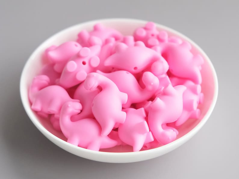 May include: A bowl filled with pink silicone dinosaur-shaped beads. The beads are all the same size and shape, and they are arranged in a random pattern.