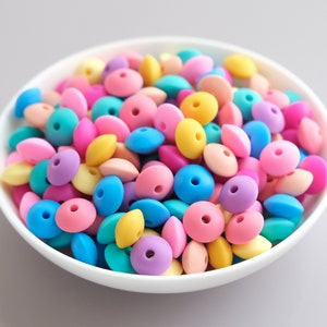 Può includere: Una ciotola piena di perline di silicone colorate. Le perline sono in varie tonalità pastello di rosa, blu, giallo e viola. Hanno la forma di ovali appiattiti con un foro al centro.