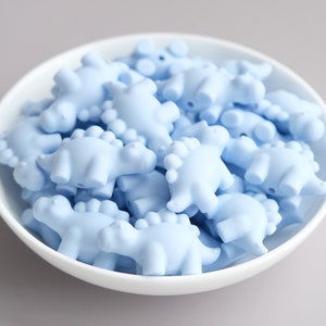 May include: A bowl filled with light blue silicone dinosaur-shaped beads. The beads are all the same size and shape, and they are arranged in a random pattern.