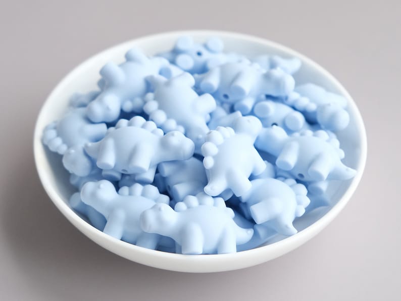 May include: A bowl filled with light blue silicone dinosaur-shaped beads. The beads are all the same size and shape, and they are arranged in a random pattern.