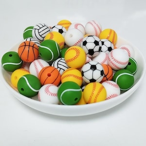 May include: A bowl filled with colourful sports-themed beads. The beads are shaped like baseballs, basketballs, footballs, tennis balls, and volleyballs. The beads are made of silicone and are perfect for making necklaces, bracelets, and other crafts.
