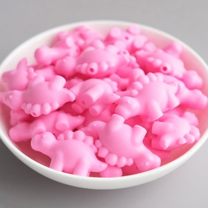 May include: A bowl filled with pink dinosaur-shaped beads. The beads are made of a smooth, rubbery material and are perfect for crafting or jewelry making.
