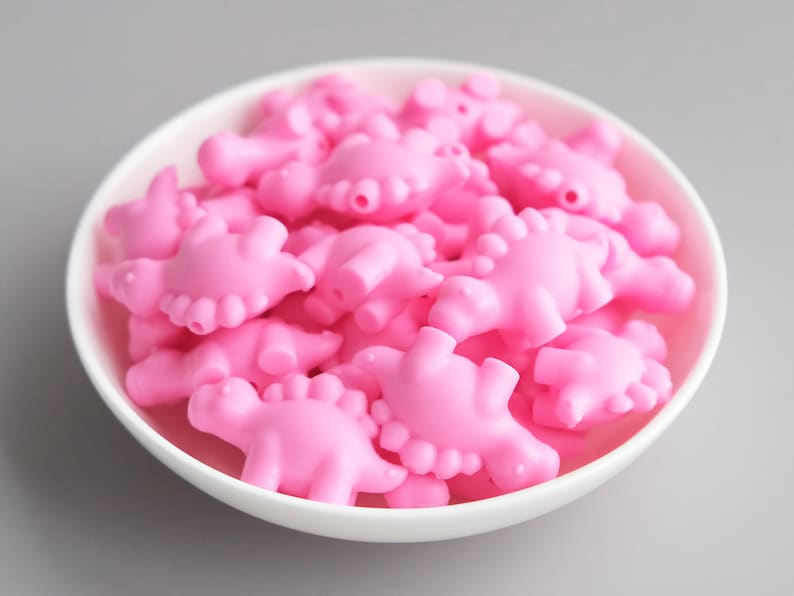 May include: A bowl filled with pink dinosaur-shaped beads. The beads are made of a smooth, rubbery material and are perfect for crafting or jewelry making.