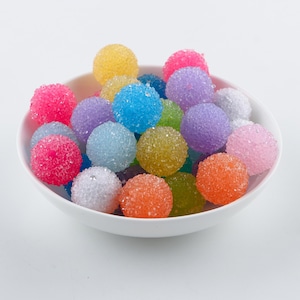 May include: A white bowl filled with colorful, sugar-coated candy balls. The candy balls are in various shades of pink, blue, yellow, green, and purple.