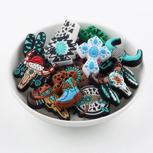 Op de afbeelding: Een verzameling kleurrijke, western-thema schoencharms met ontwerpen zoals een cowboyhoed, een laars, een schedel en een kruis. De charms zijn gemaakt van plastic en hebben een verscheidenheid aan kleuren, waaronder blauw, rood, wit en zwart.