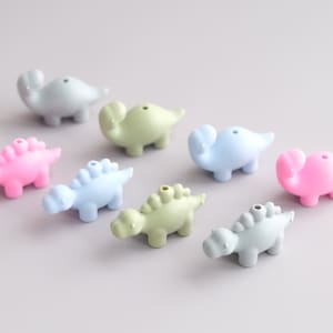 May include: A set of nine colorful dinosaur-shaped silicone beads. The beads are in shades of pink, blue, and green. They are perfect for making necklaces, bracelets, or other crafts.