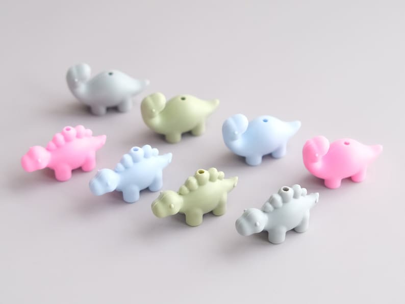 May include: A set of nine colorful dinosaur-shaped silicone beads. The beads are in shades of pink, blue, and green. They are perfect for making necklaces, bracelets, or other crafts.